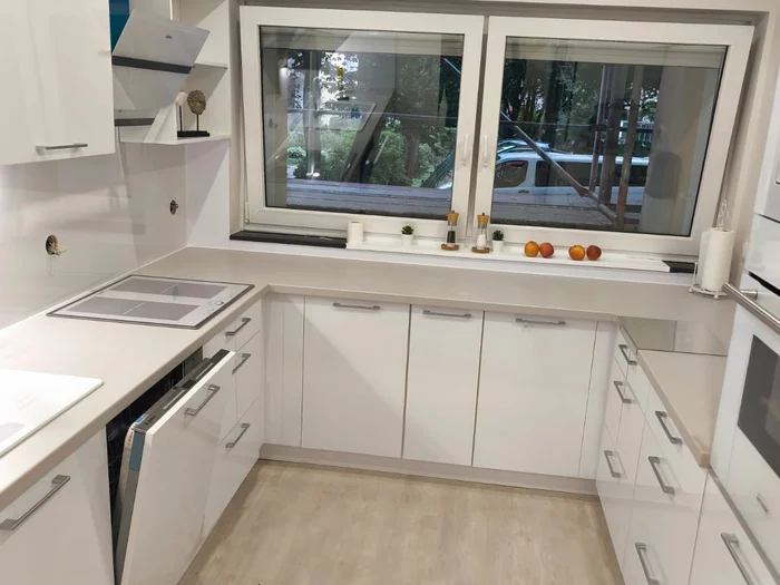 İki pencereli, açık renk tezgahlı ve pencere altında evyeli beyaz mutfak